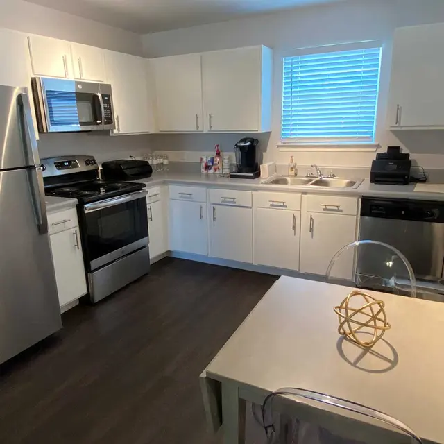 A modern kitchen featuring stainless steel appliances and white cabinetry. The space has a sleek design with a table in the foreground and a window allowing natural light in.