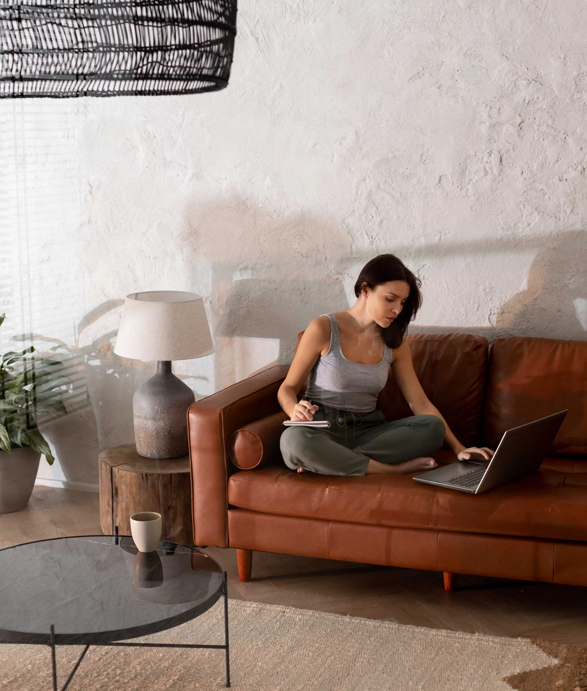 A woman sitting cross-legged on a brown leather couch, using a laptop. She has long dark hair and is wearing a gray tank top and green pants. A coffee mug is placed on a round glass table in front of her, and there are plants and a lamp in the background.