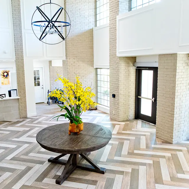 Modern lobby interior featuring a large round table with a flower arrangement, a leather bench, and tall windows allowing natural light, highlighted by a decorative chandelier.