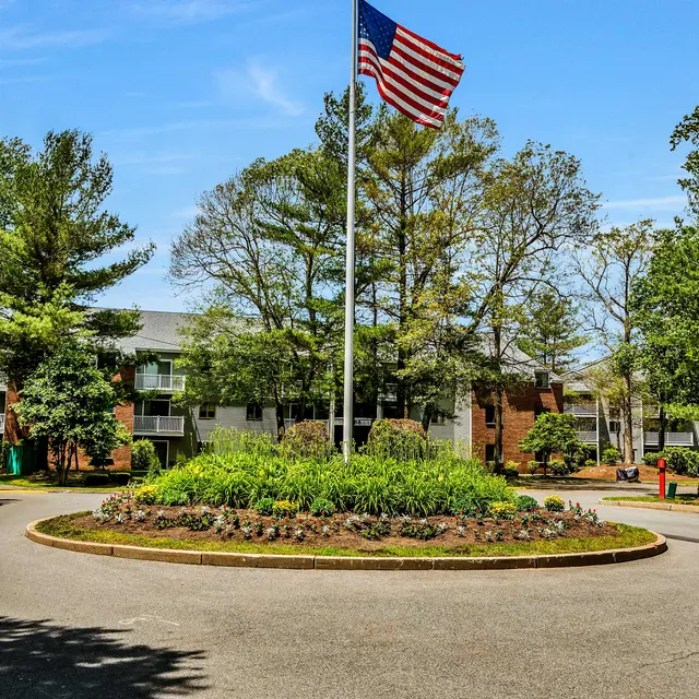 A flagpole with the American flag stands in the center of a circular flower bed surrounded by trees and a building in the background.