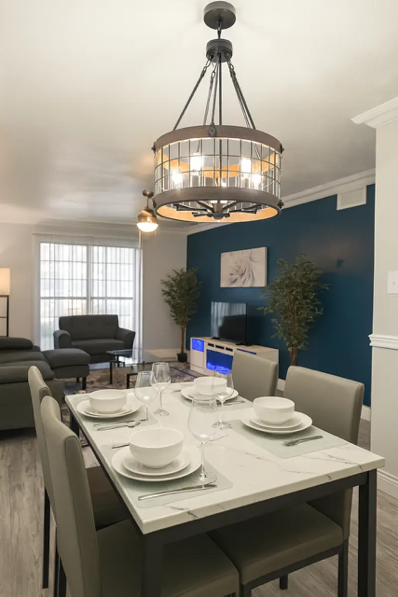 Contemporary Dining Room Interior Design A modern dining room featuring a stylish table set for dinner, with white plates and glasses. A large chandelier hangs over the table, and a cozy living area is visible in the background with a green sofa and potted plants.
