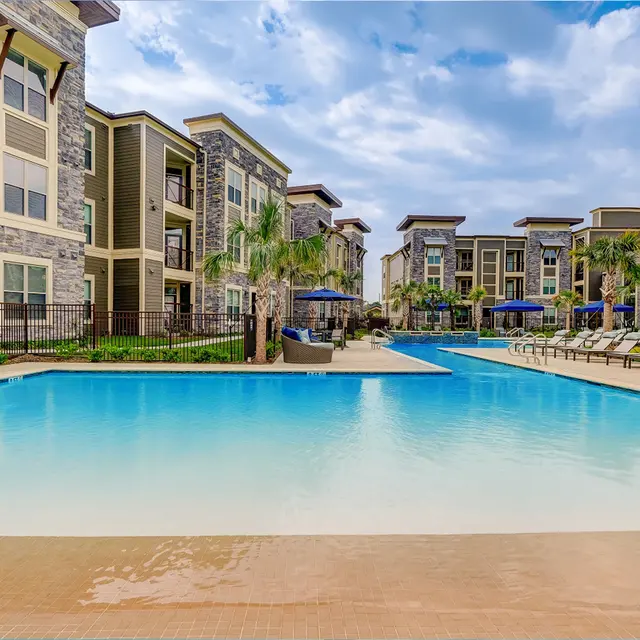 A beautiful luxury apartment complex featuring a large, calm swimming pool surrounded by lounge chairs and palm trees, under a partly cloudy sky.