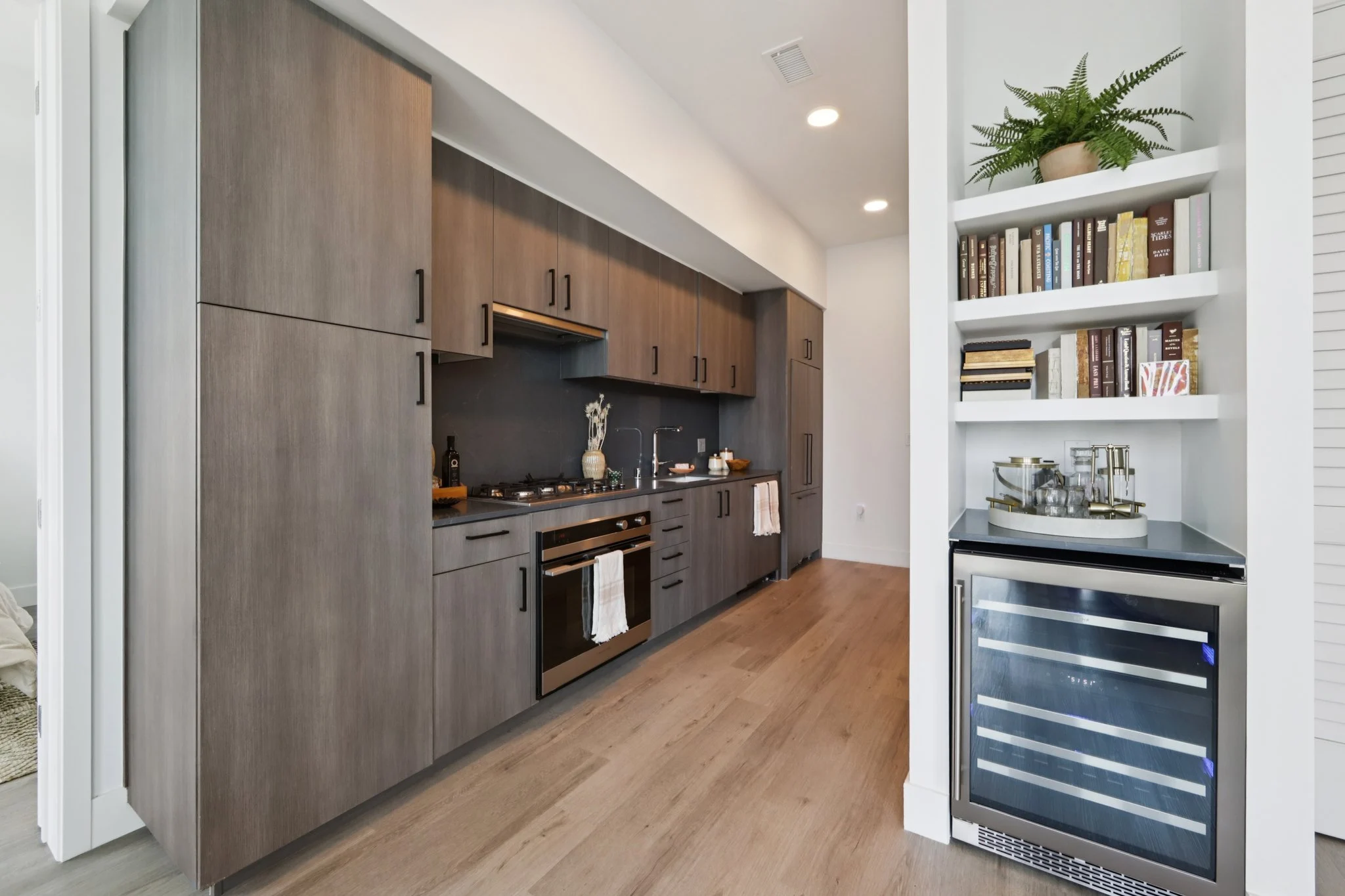 A modern kitchen featuring sleek dark cabinetry, stainless steel appliances, and a wine cooler. White walls and a minimalist open layout add to the contemporary feel.