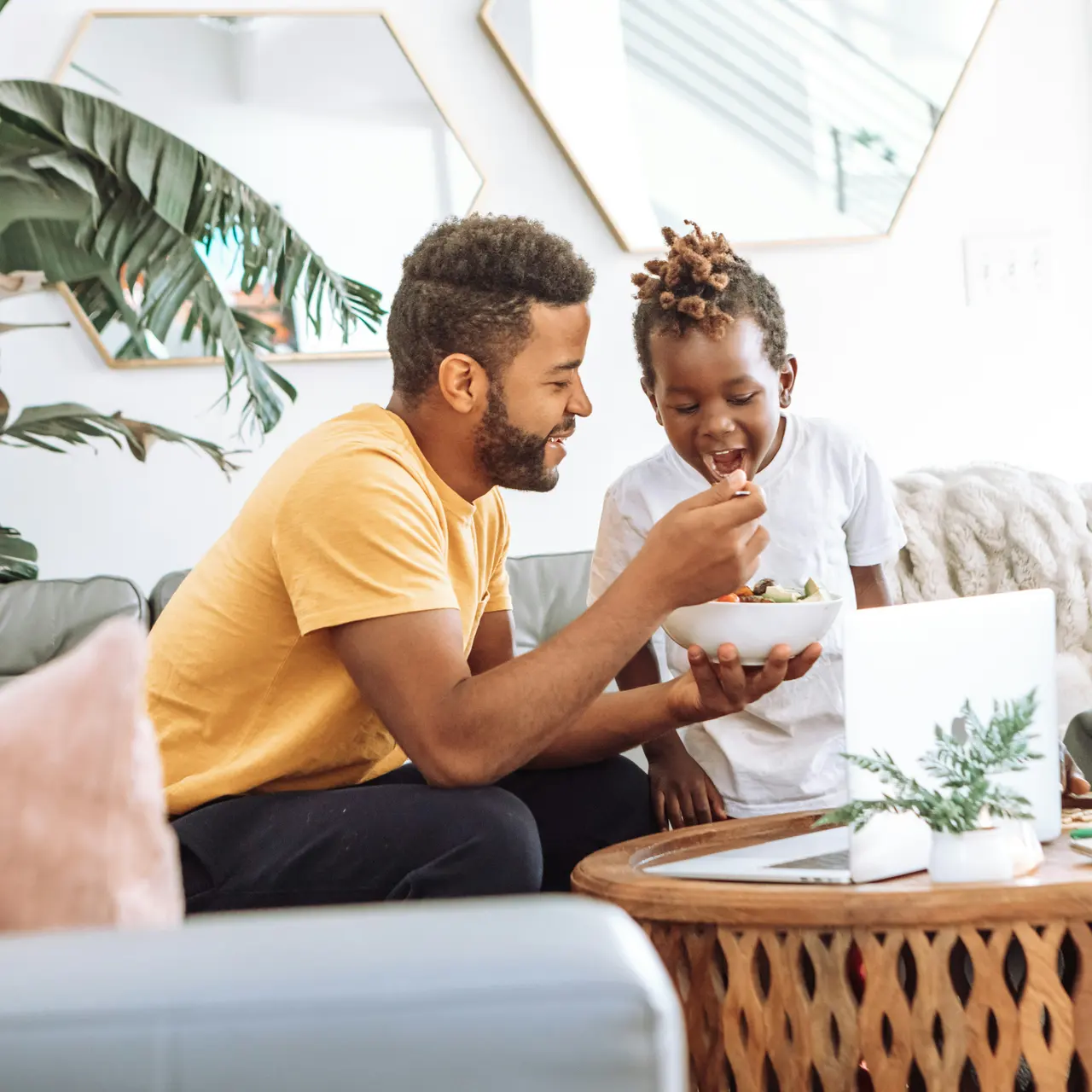 A father and son share a meal on a couch while a woman observes and smiles. The setting features greenery and a modern decor in a cozy living room.