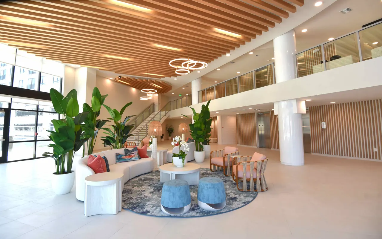 A spacious modern lobby featuring an open layout with wooden ceiling beams, ornamental plants, comfortable seating areas, and circular light fixtures.