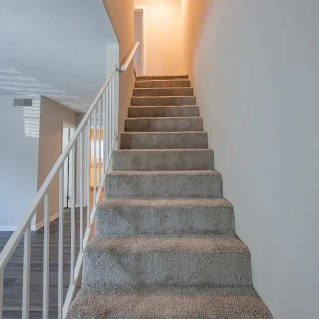 A view of a carpeted staircase leading to a brightly lit hallway at the top, with white railings on each side and soft natural light spilling in from the upper area.