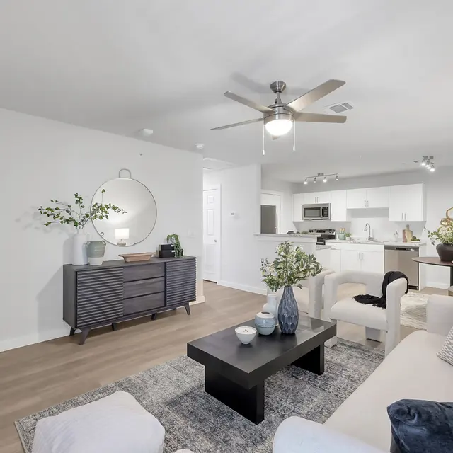 A bright, modern living room featuring a comfortable seating area with a light sofa and patterned cushions, a sleek black coffee table, and a statement circular mirror. The room has wooden flooring and large windows, allowing natural light to fill the space.