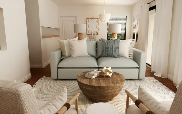 A modern living room featuring a light blue sofa adorned with decorative pillows, two beige armchairs, a round wooden coffee table, and a light rug. The room is bright with natural light, showcasing white walls and large windows with curtains.
