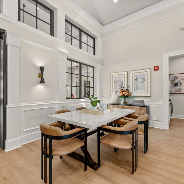A modern dining area featuring a white table surrounded by dark wooden chairs, with large windows allowing natural light in. There are decorative pieces on the table and artwork on the walls. A cozy lounge area is visible in the background.