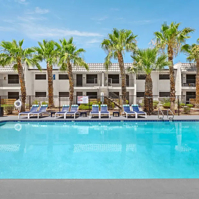 A bright and inviting swimming pool surrounded by palm trees and lounge chairs, with an apartment complex in the background.