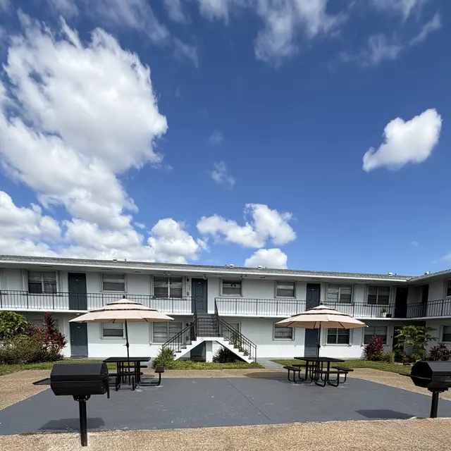 Outdoor gathering area with picnic tables, umbrellas, and barbecue grills in front of a two-story apartment building with blue sky and clouds.
