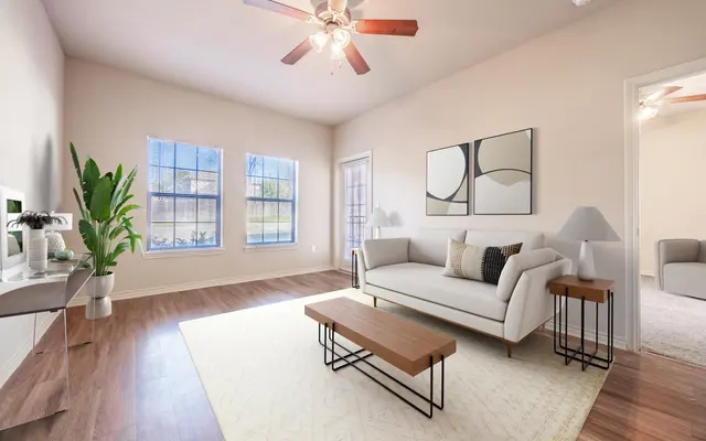 A bright and airy living room featuring large windows, a ceiling fan, a light-colored sofa with decorative cushions, two small wooden side tables, and a rug on wooden flooring. A potted plant adds a touch of greenery.