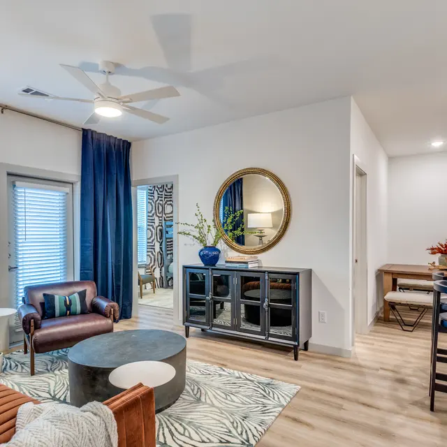 A stylish and modern living room featuring a brown leather chair, an elegant circular mirror, a coffee table, and a dining area. There are large windows with blinds allowing natural light, and decorative elements like a patterned rug and potted plants.