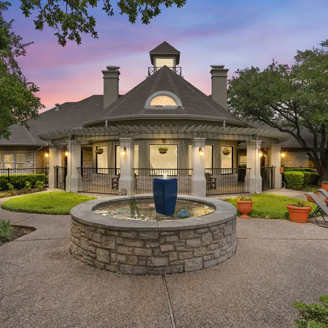 A beautifully landscaped outdoor area featuring a central stone fountain, surrounded by benches and potted plants, with a large building in the background and a colorful sky at dusk.