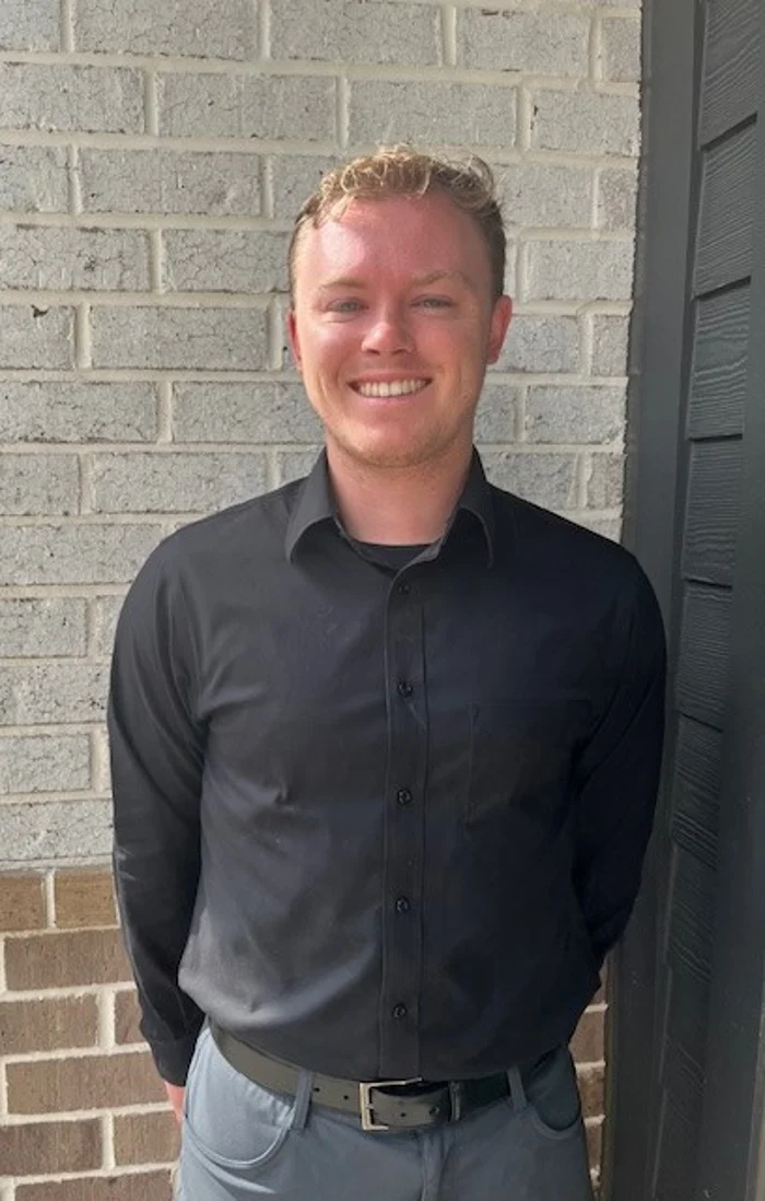 A smiling man standing in front of a brick wall wearing a black long-sleeve shirt and gray pants.