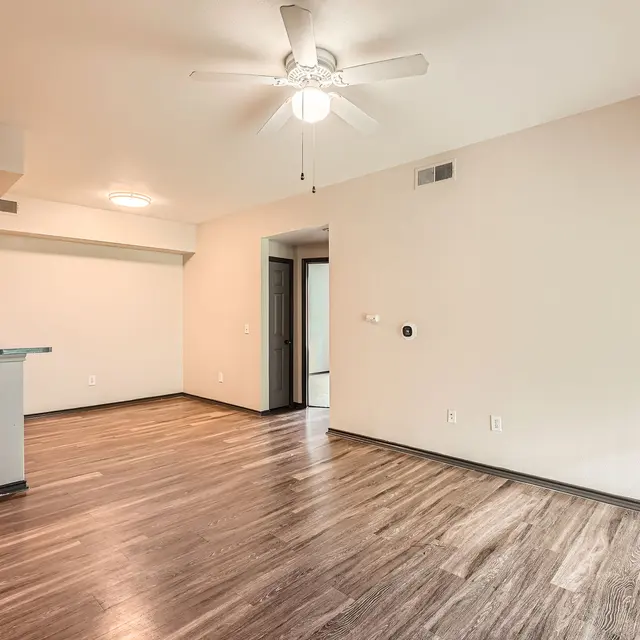 An empty living room with a ceiling fan and wood-like flooring, featuring an open kitchen area with wooden cabinets in the background.