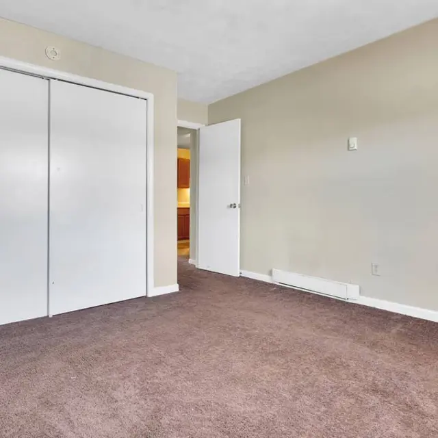 An empty room with beige walls and brown carpet, featuring a large closet with sliding white doors and a door leading to another area.