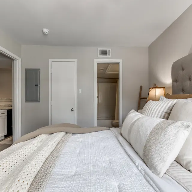 A cozy bedroom featuring a neatly made bed with a light-colored quilt and decorative pillows. The room has a neutral color scheme with soft lighting, a bedside lamp, and an open door leading to another room. The space is inviting and well-organized.