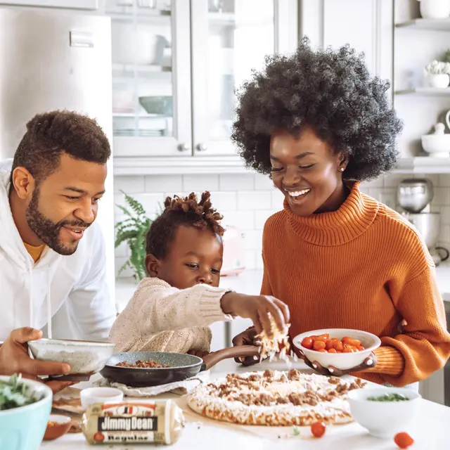 A family cooking in a modern kitchen. A man and a woman smile while a child sprinkles ingredients on a pizza crust. Fresh vegetables are on the counter.
