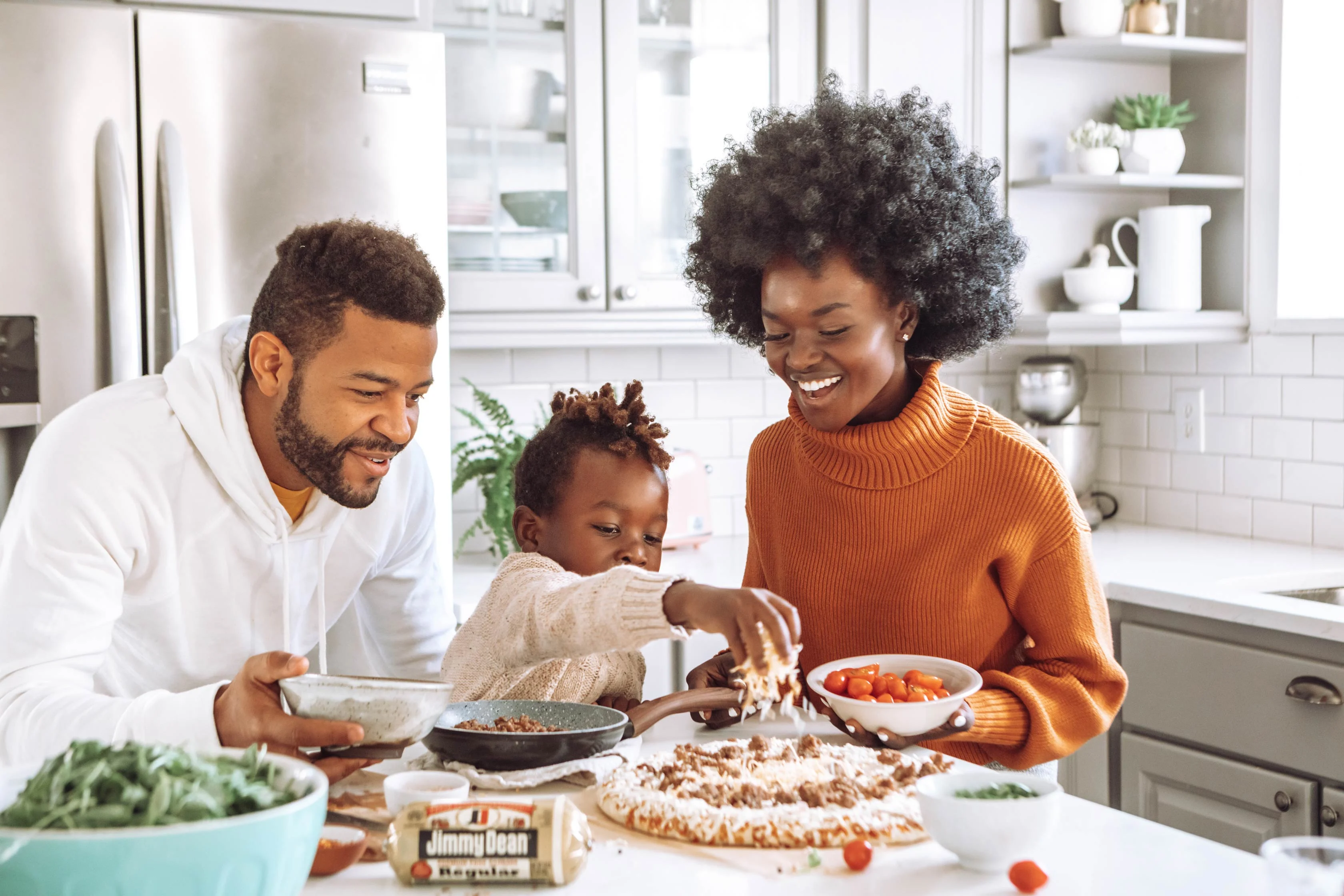 A family cooking in a modern kitchen. A man and a woman smile while a child sprinkles ingredients on a pizza crust. Fresh vegetables are on the counter.