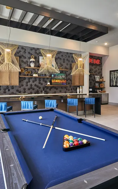 A modern game room featuring a pool table in the foreground, surrounded by tall windows that let in natural light. In the background, there is a well-designed kitchen area with wooden cabinetry and stylish lighting fixtures. A dining area with a large table and chairs is also visible, completing the inviting atmosphere.
