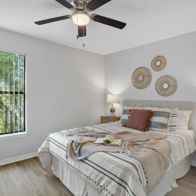 A modern bedroom featuring a queen-sized bed with striped bedding, two bedside lamps, a fan, and a window with blinds, allowing natural light to enter.