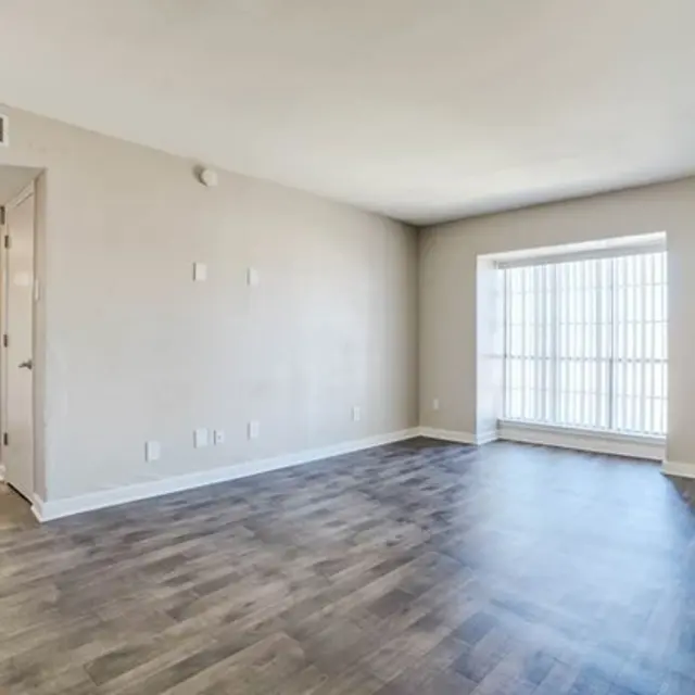 A spacious, empty living room with light-colored walls and dark wooden flooring, featuring a large window allowing natural light to enter.