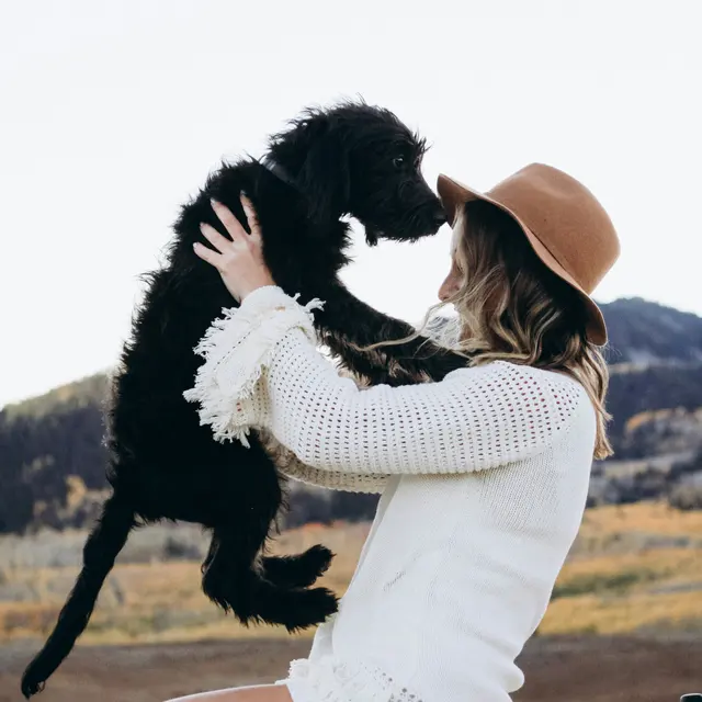 A woman dressed in a white sweater and a hat, sitting on the roof of a car, holding a black puppy close to her face against a picturesque landscape.