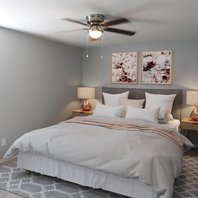 A modern bedroom featuring a plush bed with decorative pillows, bedside tables with lamps, and a light gray color scheme. The room has a ceiling fan and framed art on the wall.