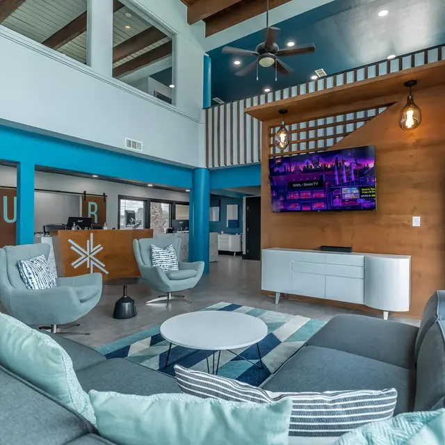 A modern lobby interior featuring a stylish gray sofa, two light blue chairs, a round coffee table, and a large television mounted on a wooden wall. The space has high ceilings with wooden beams and vibrant blue walls, adding a contemporary feel.