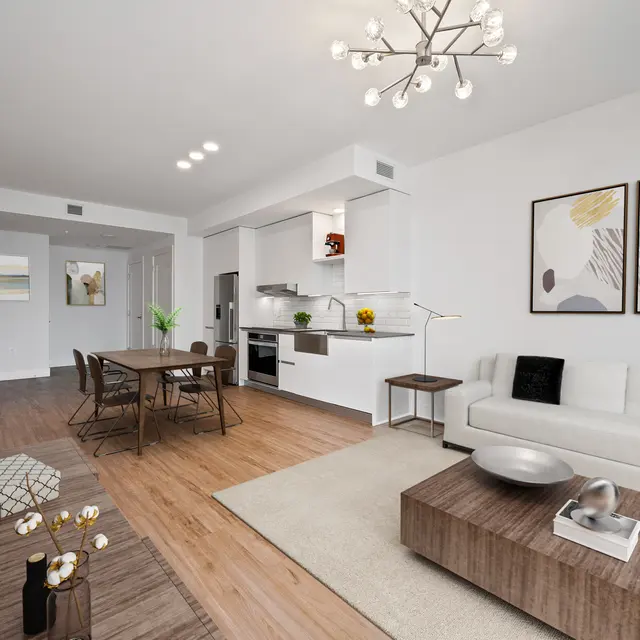 A modern living space featuring a white sofa, a wooden coffee table, a dining area with wooden chairs, and a stylish chandelier. The kitchen is open and equipped with contemporary appliances. Abstract art hangs on the walls.