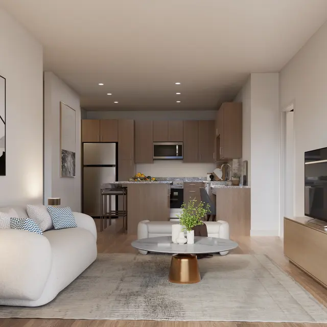 A modern living room featuring a light-colored sofa, coffee table, and a stylish entertainment unit with a TV. The room has a cozy atmosphere with hardwood floors and contemporary decor elements like a plant and artwork on the walls. The kitchen area is visible in the background with wooden cabinetry and modern appliances.