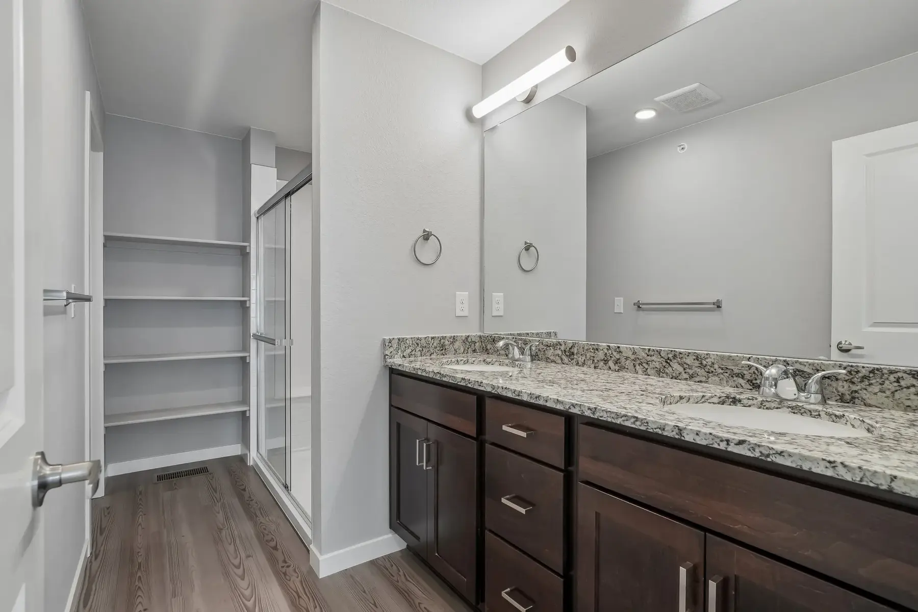 Contemporary Bathroom Design A modern bathroom featuring a double vanity with dark wooden cabinetry, granite countertops, a large mirror with integrated lighting, a glass shower enclosure, and shelving. The floor is adorned with elegant wood-like tiles.