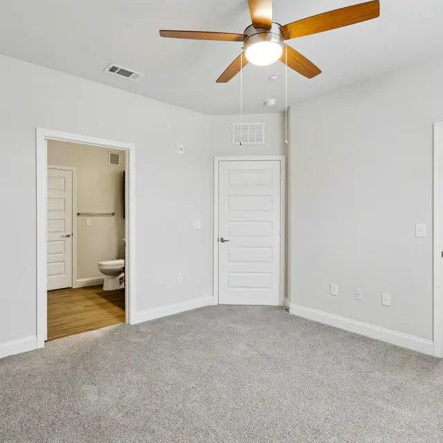 A spacious empty room with light gray carpet, a ceiling fan, and two doorways leading to a bathroom and a closet.