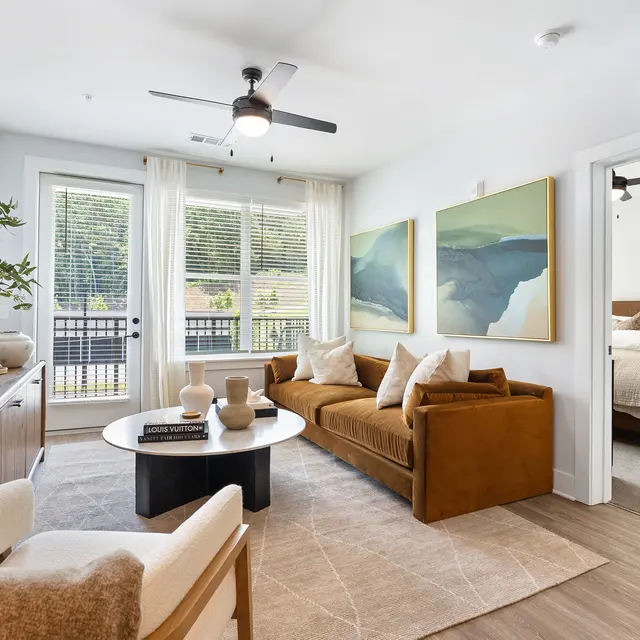 A modern living room featuring a brown sofa, light-colored armchair, round coffee table, and large artworks on the wall. Large windows allow natural light to fill the space, with a view of greenery outside. The adjacent room, partially visible, is styled with beige bedding.