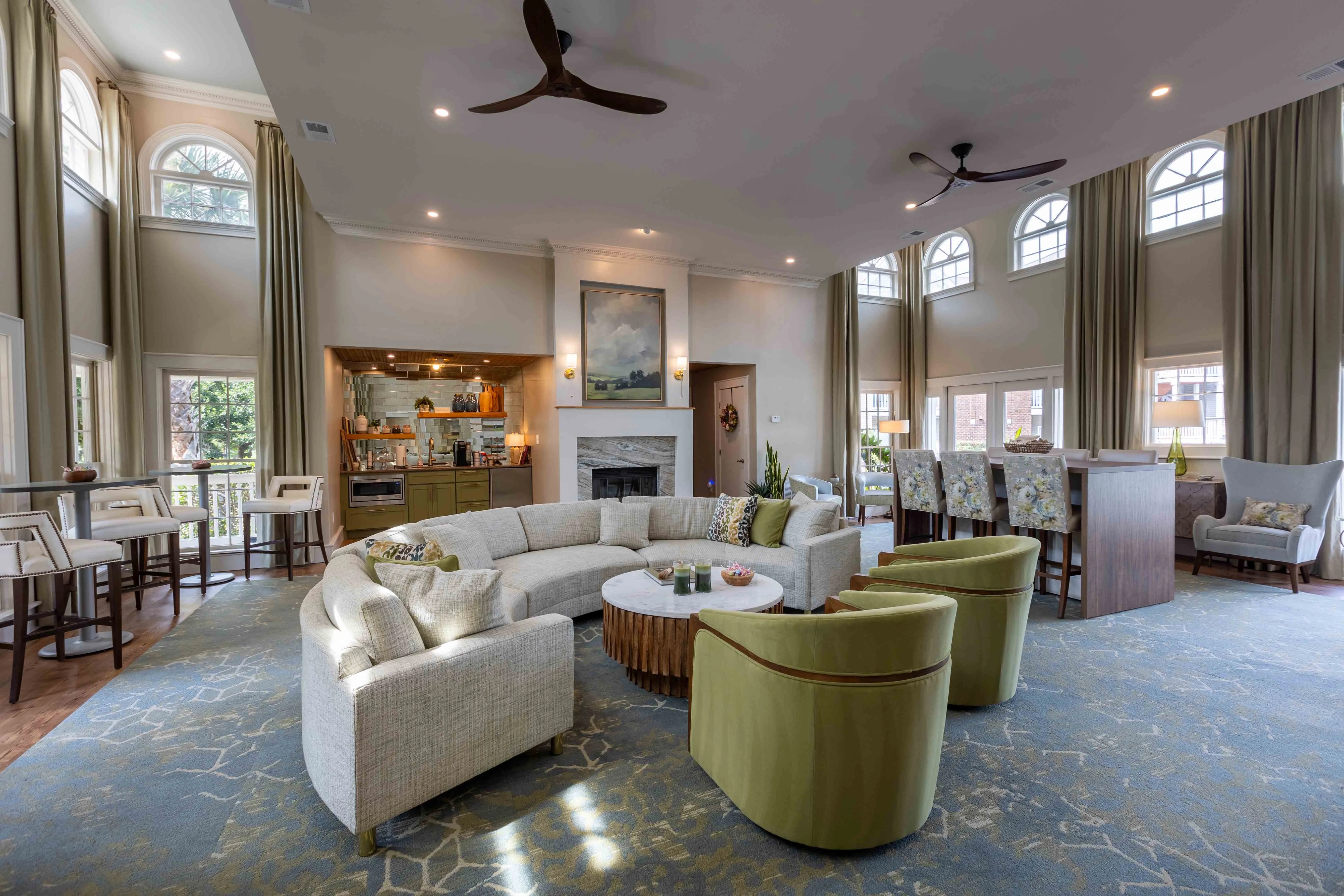 A spacious lounge area with large windows, featuring a circular light-colored sofa, green chairs, a round wooden table, and a kitchenette in the background.