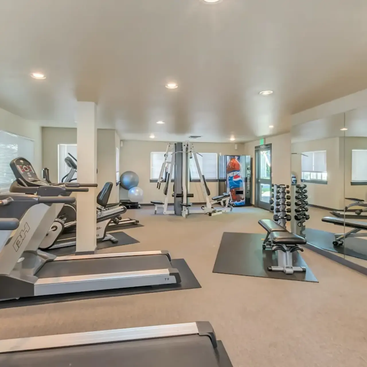 A spacious exercise room featuring various fitness equipment including treadmills, stationary bikes, weight benches, and a multi-gym apparatus. The room is well-lit with mirrors on the walls and has a clean, modern design.