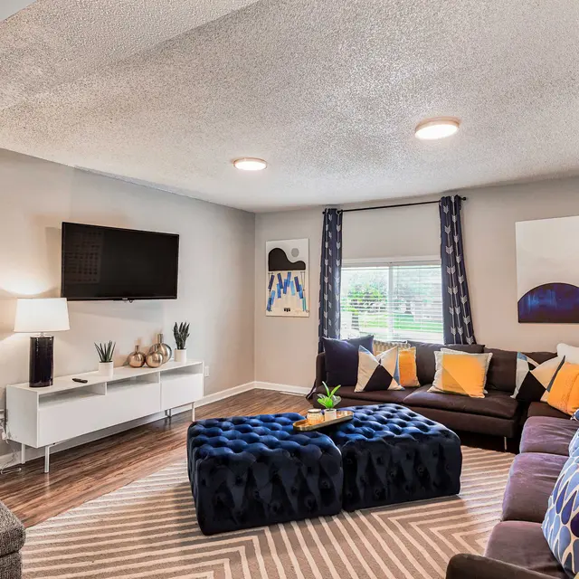 A cozy modern living room featuring a large sectional sofa, an ottoman, a wall-mounted TV, and decorative art on the walls. Natural light enters through a window adorned with patterned curtains.