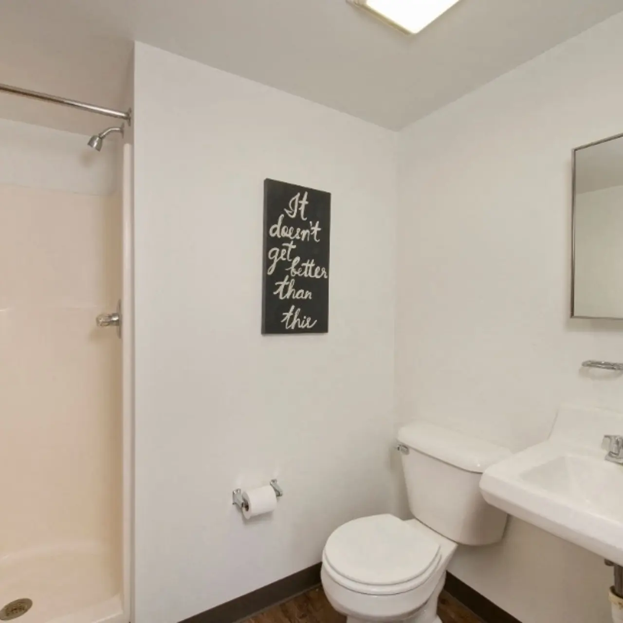 A modern bathroom featuring a shower stall, toilet, and sink with a rectangular mirror above. A motivational quote is displayed on the wall.