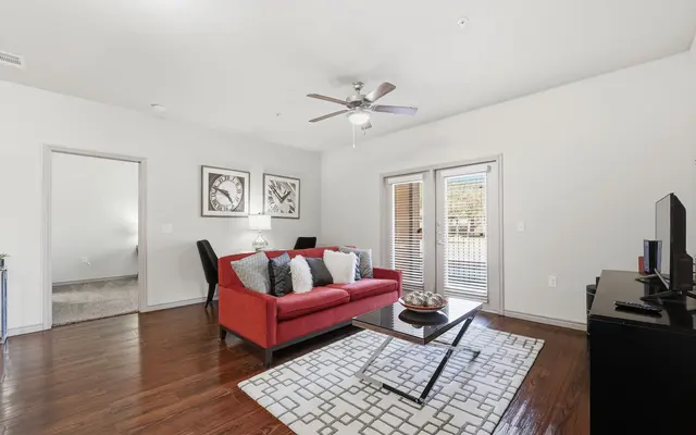 A modern living room featuring a red sofa, a patterned area rug, and a coffee table. There are two pieces of artwork on the wall and sliding glass doors leading to an outdoor space. A ceiling fan hangs from the ceiling, and a TV stand is visible on the right.