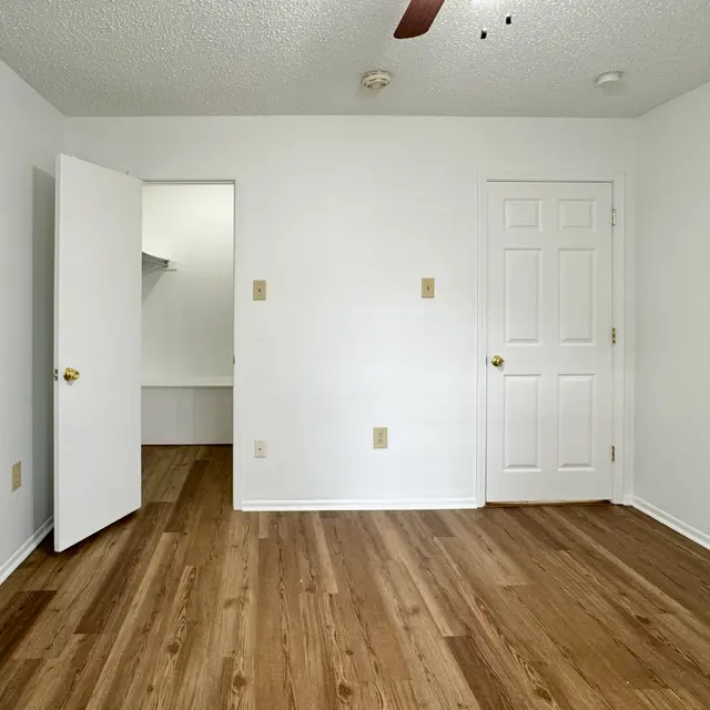 A vacant room showcasing a light-colored wall, a wooden floor, and two doors—one leading to a closet and the other to an adjoining space. A ceiling fan is visible, and the overall atmosphere is bright and spacious.