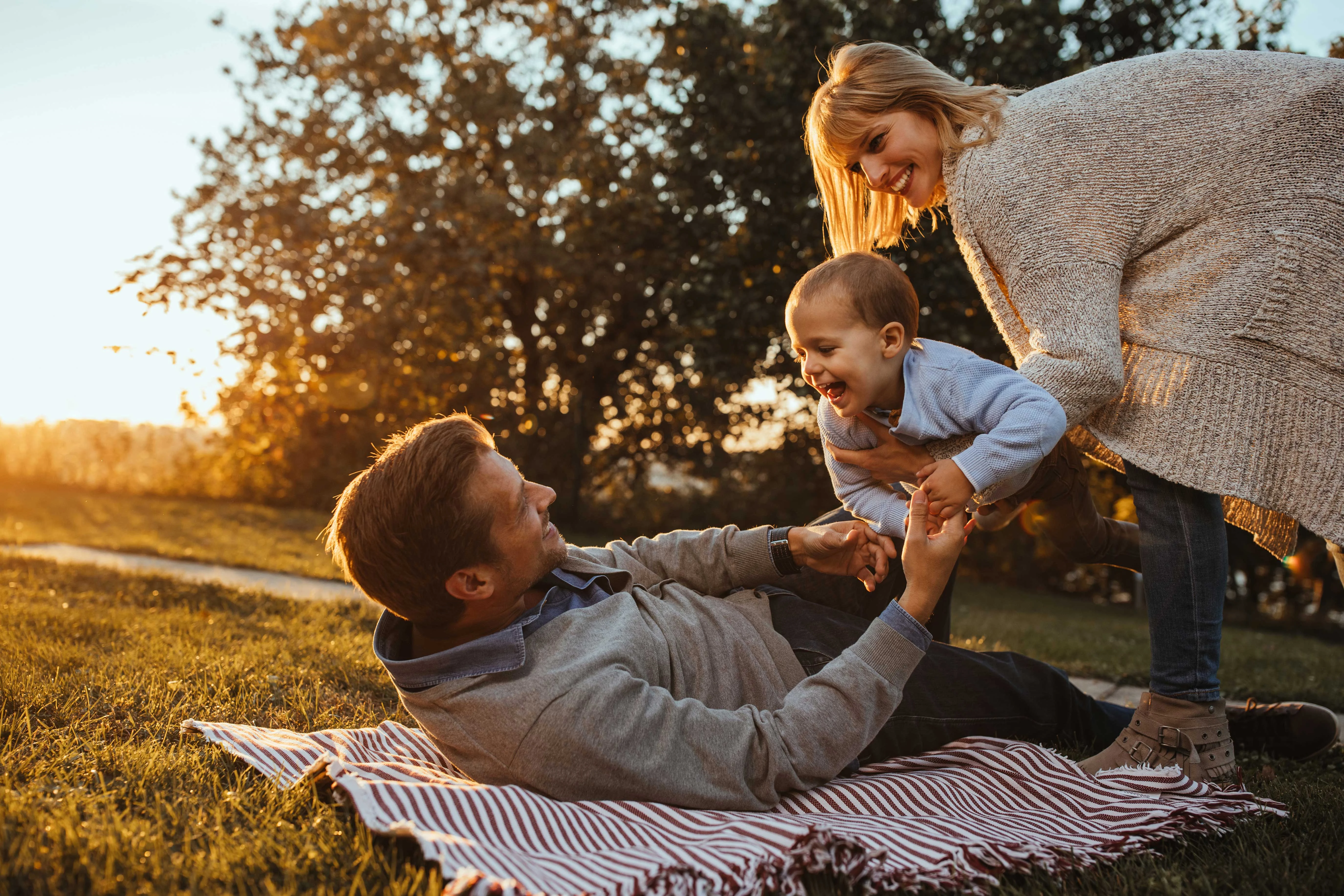 A family enjoying time outdoors during sunset. A father lies on a blanket, holding his young child, while the mother stands nearby, watching and smiling. The scene is filled with warm sunlight and greenery.