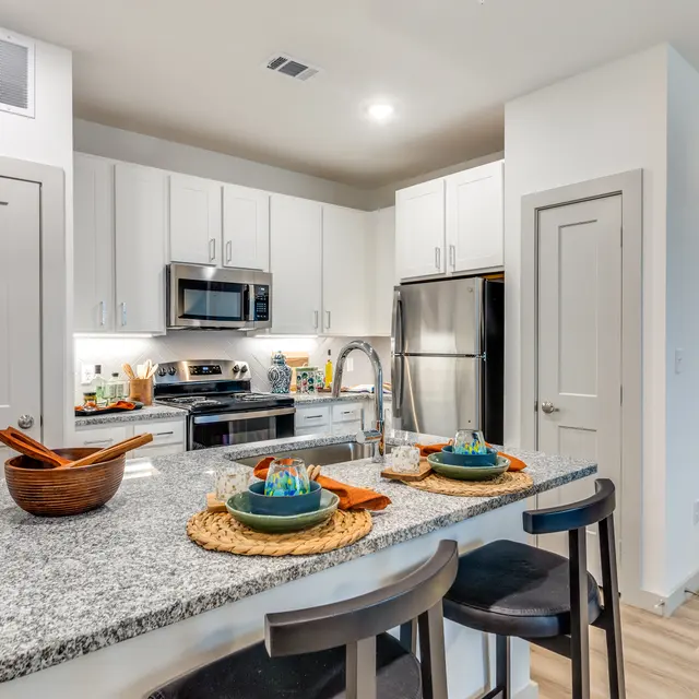 A modern kitchen featuring a large island with granite countertop, wooden bowls, and plates. It includes stainless steel appliances and white cabinetry, with a sleek design and warm lighting.
