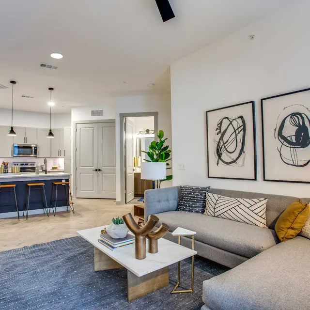 A modern living room featuring a gray sectional sofa, a coffee table with decorative items, and wall art. The kitchen area is visible in the background, showcasing stainless steel appliances and a breakfast bar with stools.