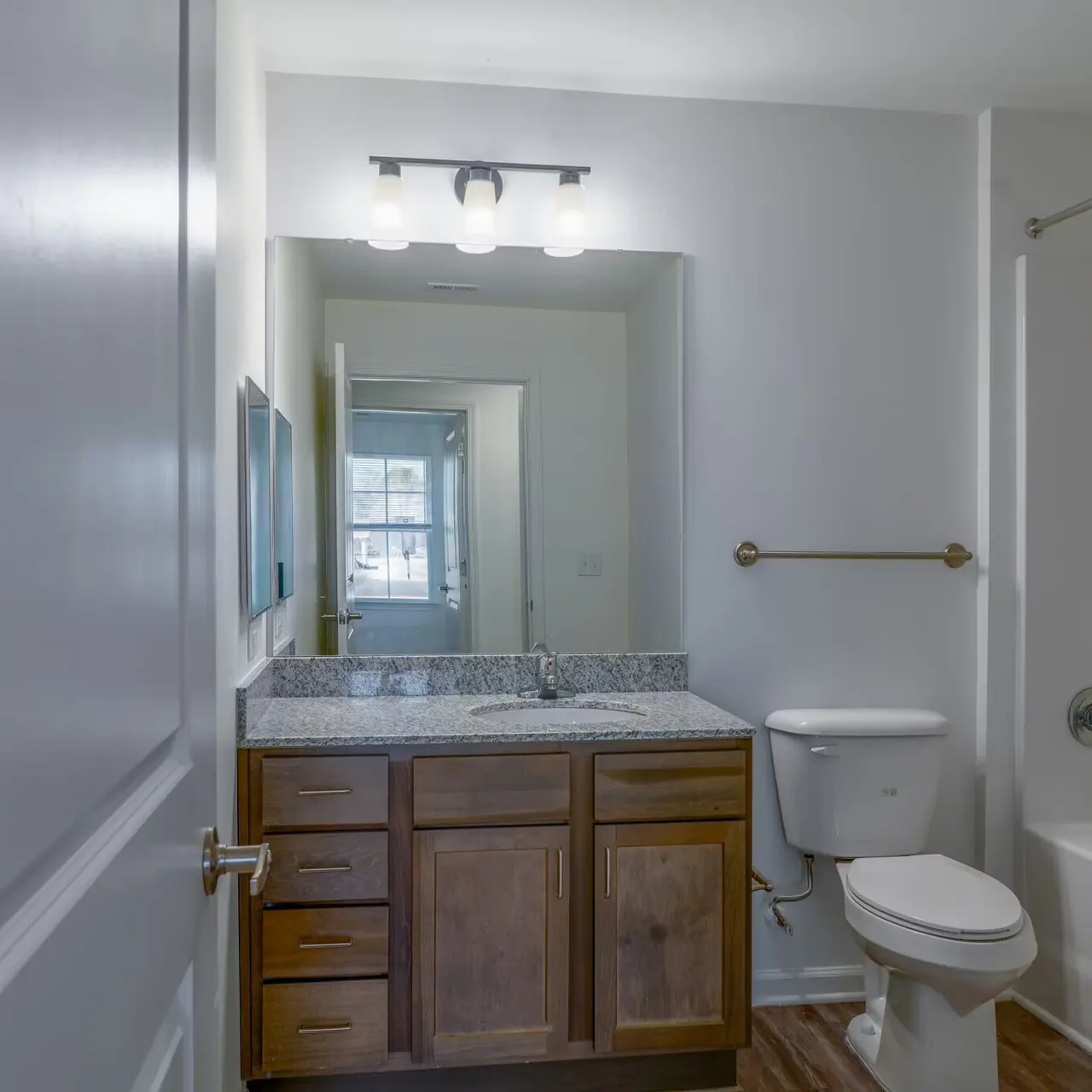 A modern bathroom featuring a brown wooden vanity with a granite countertop, a white toilet, a white bathtub with a shower, and a large mirror above the sink. The walls are painted white and the flooring is wooden.