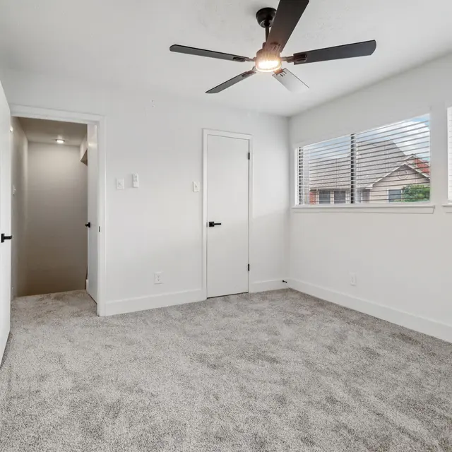 A bright, modern bedroom with light grey carpeting and a ceiling fan. It features two windows with blinds, a door leading to a hallway, and a doorway leading to an ensuite bathroom.