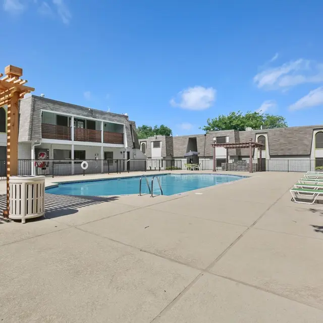 A pool area in an apartment complex featuring a swimming pool, lounge chairs, and a pergola. The building has multiple stories with balconies, surrounded by a fenced area.