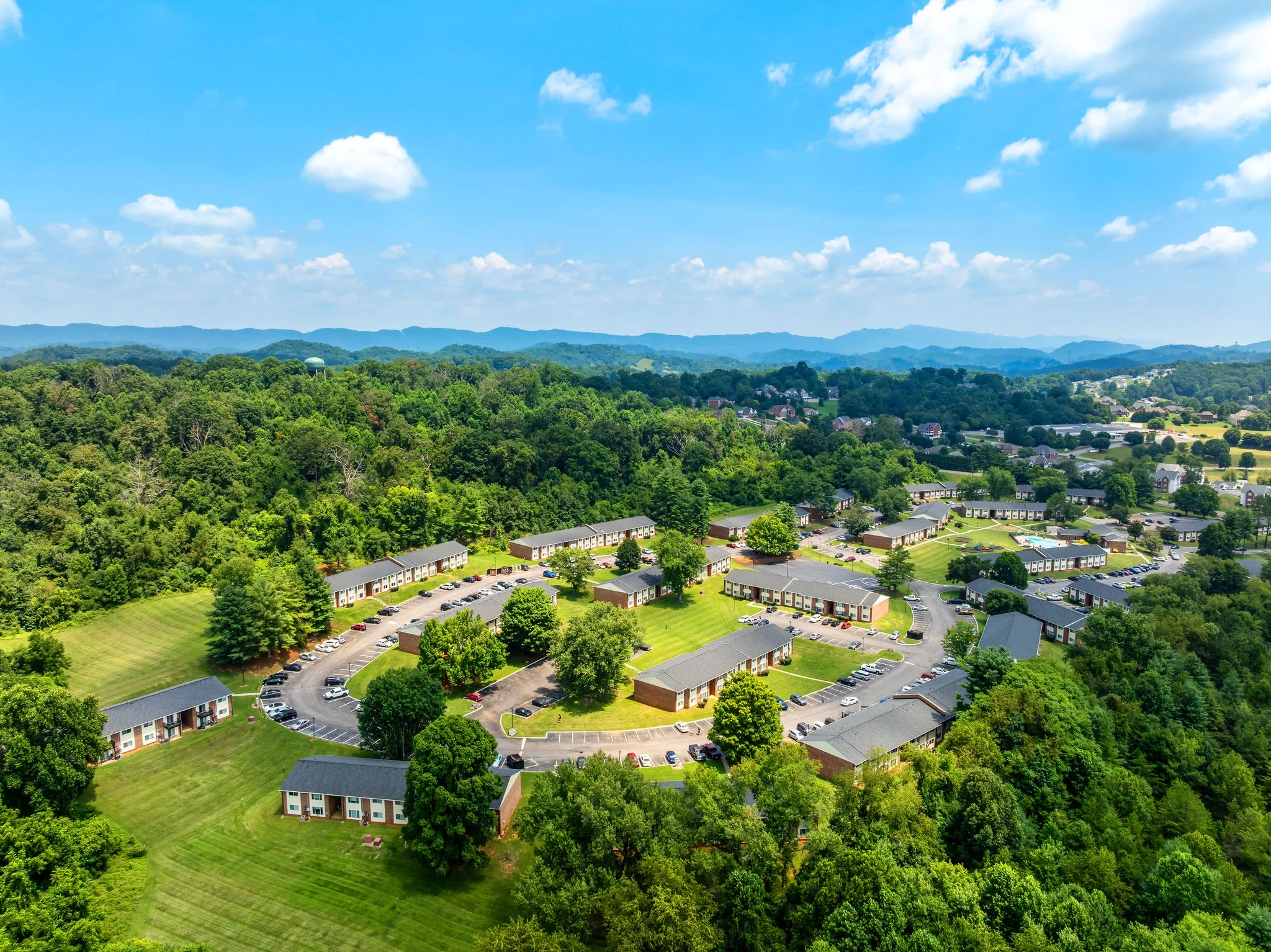 Aerial view of an apartment community surrounded by green hills and trees under a clear blue sky.