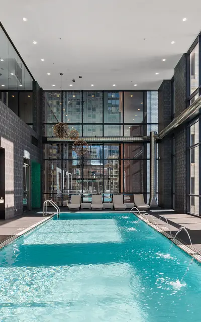 A modern indoor swimming pool surrounded by large windows, showcasing a city view. The pool area features sleek design elements and comfortable lounge chairs.