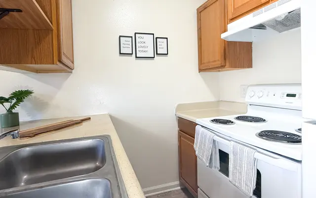 A cozy kitchen featuring light-colored walls, wooden cabinets, and a clean countertop with a double sink. The stove is white, with three burners visible. Above the countertop, there are three framed quotes on the wall.